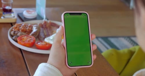 Woman Holding Smart Phone with Green Screen Chroma Key at Cafe Table with Filled with Food