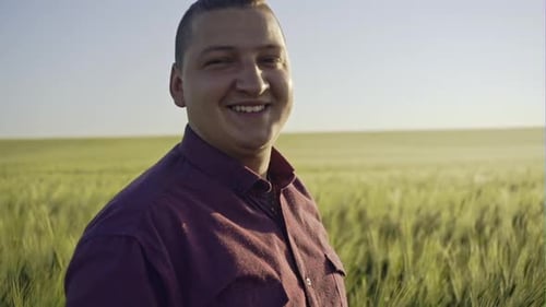 Close Up Portrait of Smiling Man Standing in Green Wheat Field on Sunset and Looking at the Camera