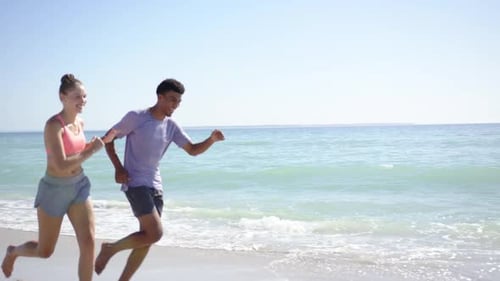 A young Caucasian woman and a biracial man, dressed in athletic wear, are jogging along the beach
