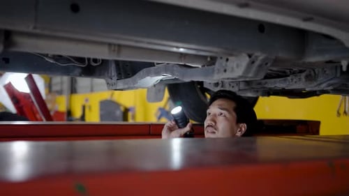 Young asian mechanic with light in hands looking at steering chassis and suspension under a car