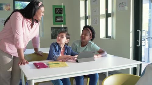 African American teacher walking left, leaning and pointing at tablet, coaching two teenage girls