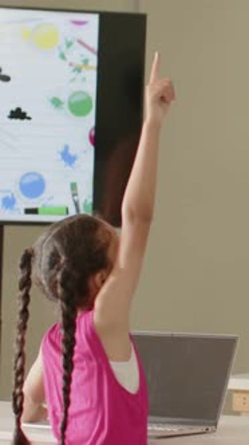 Responding to monitor, female student in pink top raising right arm and pointing, with laptop