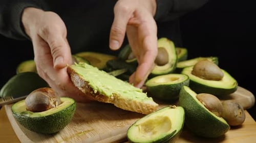 A man spreads avocado flesh on bread and showcases the toast in a close-up shot. Avocado halves.