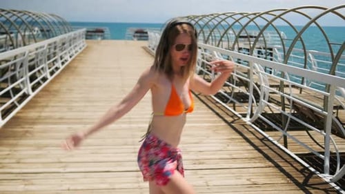 A Woman in orange swimsuit dances on pier with blue sky and sea landscape on background