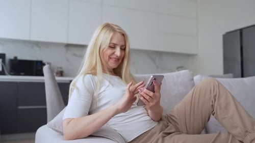 Woman Sits on a Modern Couch in a Living Room Happily Using a Smartphone for Relaxation