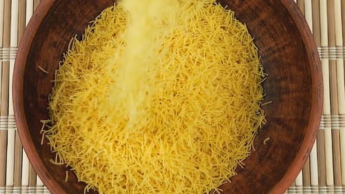 Thin small pasta is poured into a brown ceramic bowl against a bamboo mat. Top view. Slow motion.