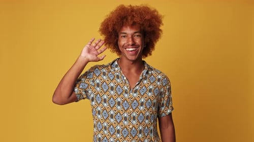 Happy guy waving palm in greeting gesture isolated on yellow background in studio