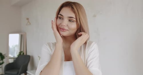 Blonde woman in robe gently touching face indoors