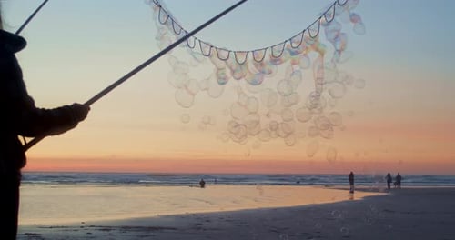 Von einer Person mit einem Seifenblasenstab schweben Blasen über den Strandhimmel bei Sonnenuntergang