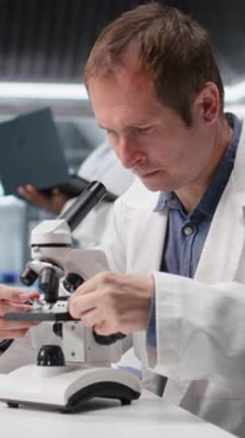 Funcionário de vídeo vertical estudando bandeja de amostra sob o microscópio para estudo de pesquisa