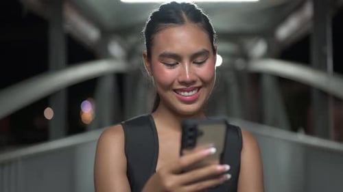 Young Asian woman at night using mobile phone in city walking bridge