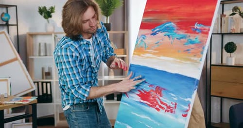 Modern man artist creating picture applying paints on picture with his hands in home workshop.