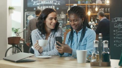 Two Beautiful Girlfriends Sitting in the Coffee Shop and Chatting, One Shows Pictures on Her Smartp