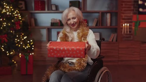 Glückliche attraktive Seniorin im Rollstuhl, die der Kamera ein Geschenk hält und zeigt, Kaukasierin
