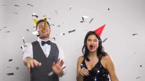 Happy stylish couple celebrating holidays with blowers and confetti indoors