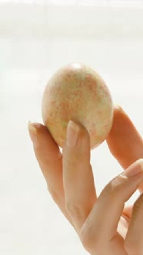 Vertical video. Hand holding a pastel-colored Easter egg up to the light, highlighting the intricate
