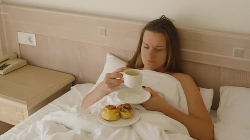 White bed in the room and the woman in it drinks coffee, sunlight illuminates the black bra.