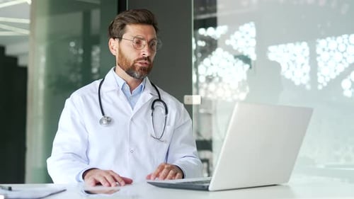 Confident doctor talking on video call using laptop in hospital clinic. Male medical worker