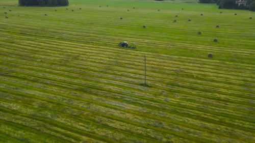 Aerial rotating view over hay baling tractor on a green farmland field with hay rolls at summer day