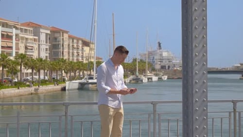 a happy young man takes selfies on a metal bridge with passing cars, in a sunny city by the sea, wit