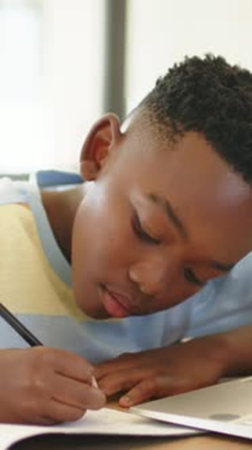 Vertical video: Starting homework, boy in striped shirt writing at home, glancing right with pencil
