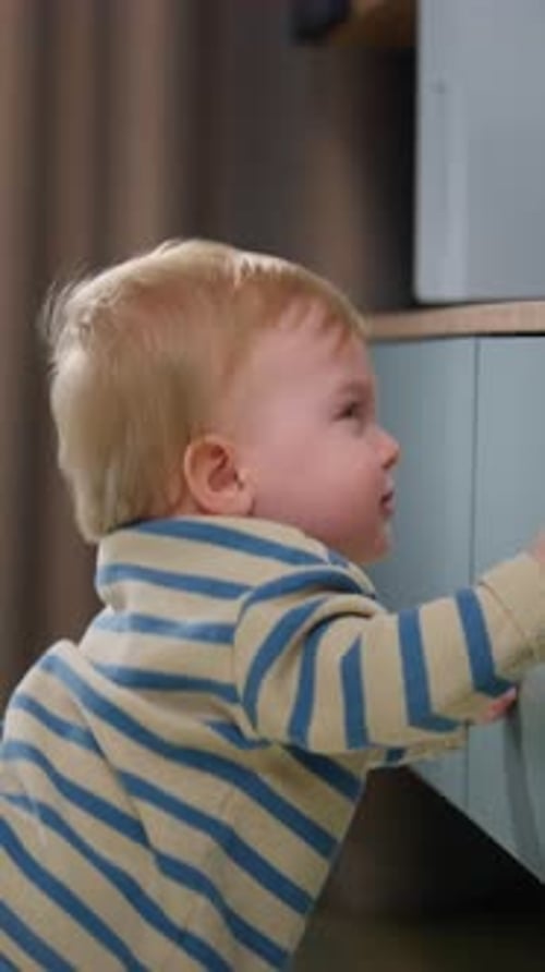 Toddler Standing at Right Foreground Gripping Low Cabinet During Hesitant Standing Transition