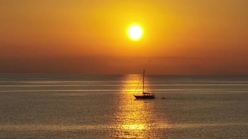 Side view of backlit sailboat silhouette floating on open ocean in sun ray beam under orange golden
