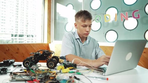 Teen Boy Codes Laptop Next to Robot Car