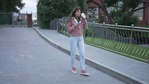 Young Woman Strolls Along the Street Sipping Coffee and Checking Her Smartphone Dressed in Casual