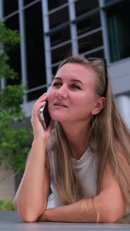 Eine junge Frau lehnt sich an einen Tisch im Freien und spricht auf ihrem Smartphone vor einem Glasbüro