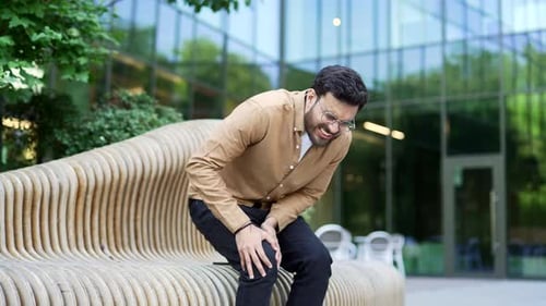 Handsome man suffers from painful severe knee joint pain sitting on a bench on the street near