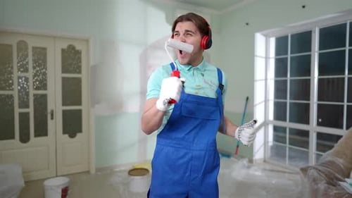 Portrait of Joyful Young Man with Paint Roller in Headphones Singing Dancing Standing in Living Room