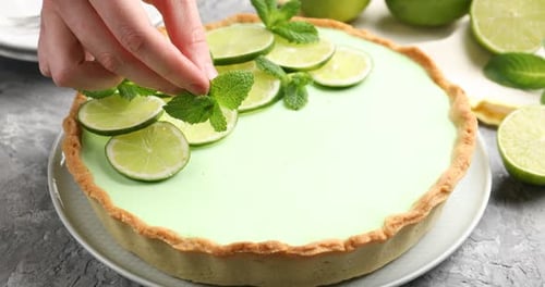 Frau legt Minzblätter auf Limettenkuchen am grau strukturierten Tisch, Nahaufnahme