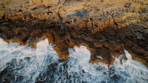 Woman stands on the rugged coastline rocks with foamy ocean waves at sunset