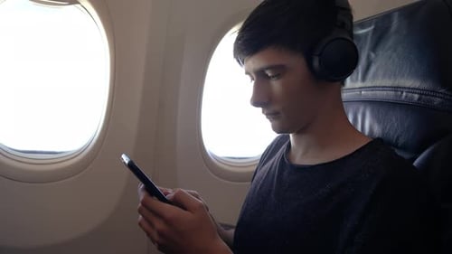 Teenage Boy Enjoying Smartphone on Flight While Wearing Headphones By Airplane Window