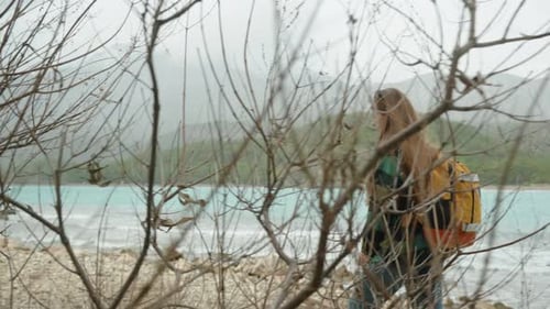 A female tourist with a backpack walks along the rocky shore with turquoise sea water and mountains