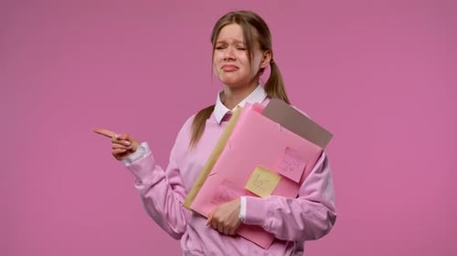 A Young Female Student Points Disappointedly to the Side Holding Folders and Notes in Her Hands