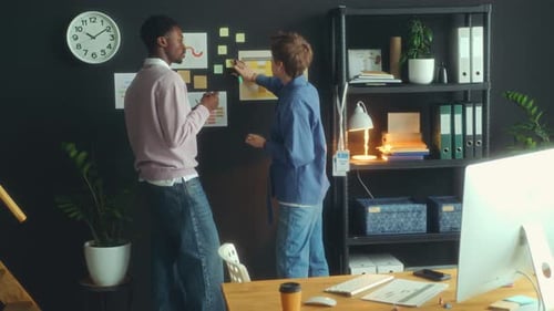 Two Coworkers Putting Sticky Notes on Office Wall and Discussing Ideas