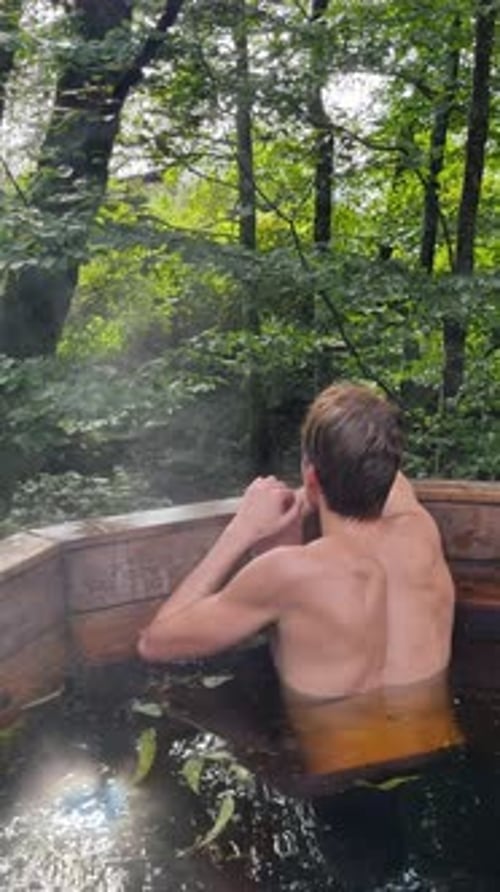 Homem relaxando na banheira de hidromassagem de madeira ao ar livre cercado por uma floresta exuberante Vídeo vertical