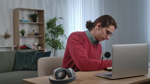 Young Adult Man Spending Time at Home Man Student Sits at Desk Works on Laptop Talking Online By