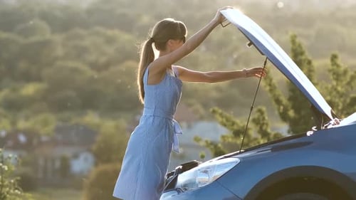 Puzzled Young Female Driver Standing Near a Broken Car with Open Up Hood Inspecting Her Vehicle