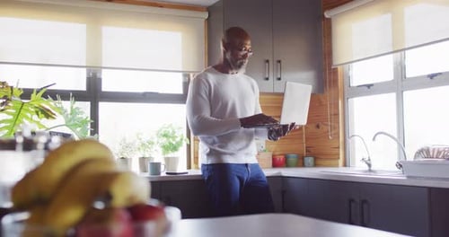 Happy senior african american man in log cabin, standing in kitchen and using laptop, slow motion
