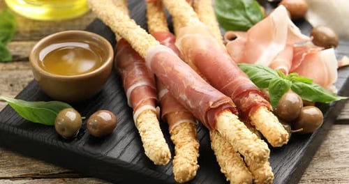 Woman taking grissini breadstick with prosciutto from board at wooden table, closeup