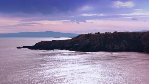Approaching the rocky shoreline at sunset time. Gleaming waterscape reflect pink sky.