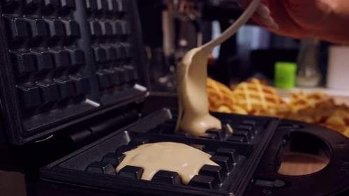 Close Up Of Batter Being Poured Into Waffle Maker With Finished Waffles Visible Behind, Detailed
