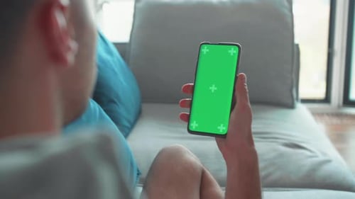 Young Caucasian Man at Home Lying on a Couch Using Smartphone with Green Mockup Screen Doing Swiping