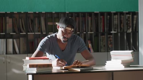 Focused Student Writing at Library Table Surrounded By Books
