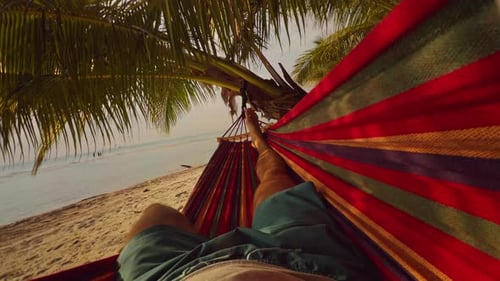 First Person View Of Man Rocking In Hammock Under Palm Trees With Colorful Fabric And Quiet Ocean At