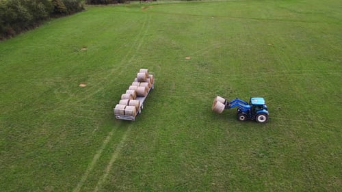 A blue tractor is seen loading large round hay bales onto a trailer in a vast green field. The foota
