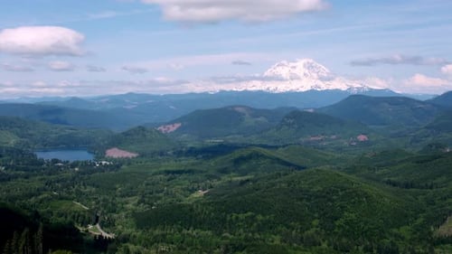 Snow-capped Mount Rainier towers above rolling forested hills and a scenic lake under a partly